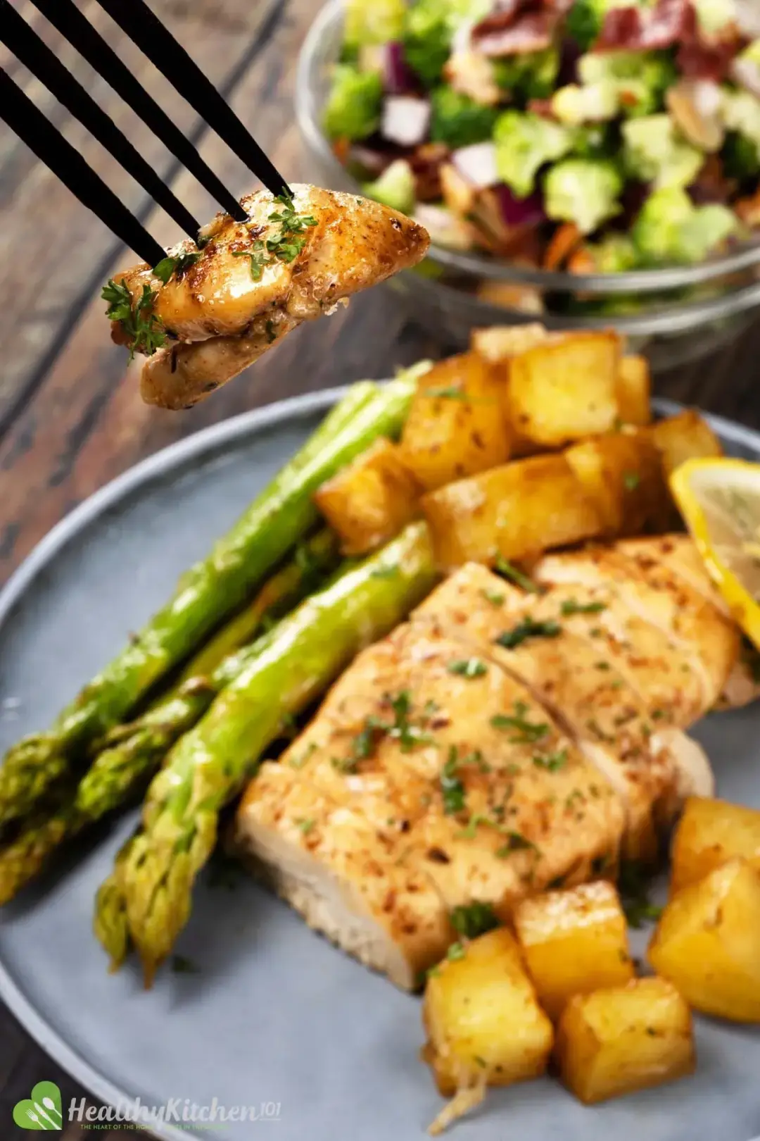 A piece of chicken held by a fork with a plate containing asparagus, potato cubes, and a piece of chicken breast in the back