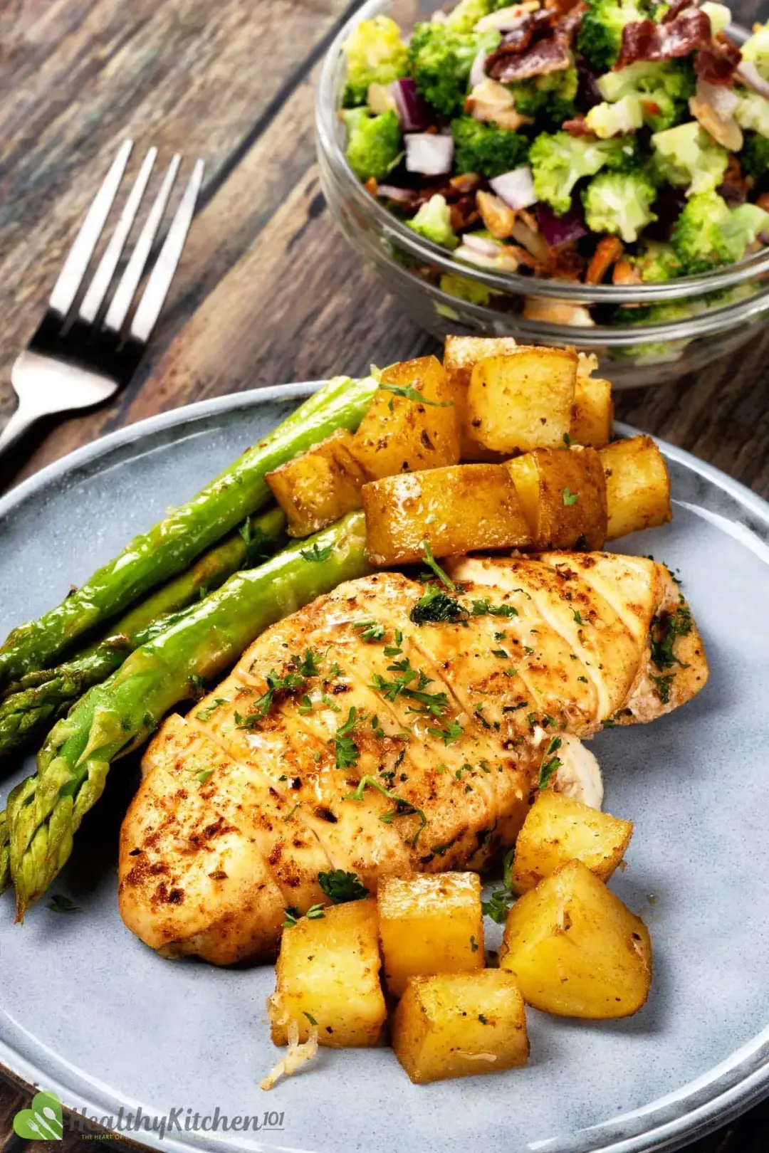 A plate of chicken breast, potato cubes, and asparagus stalks next to a small bowl of broccoli salad