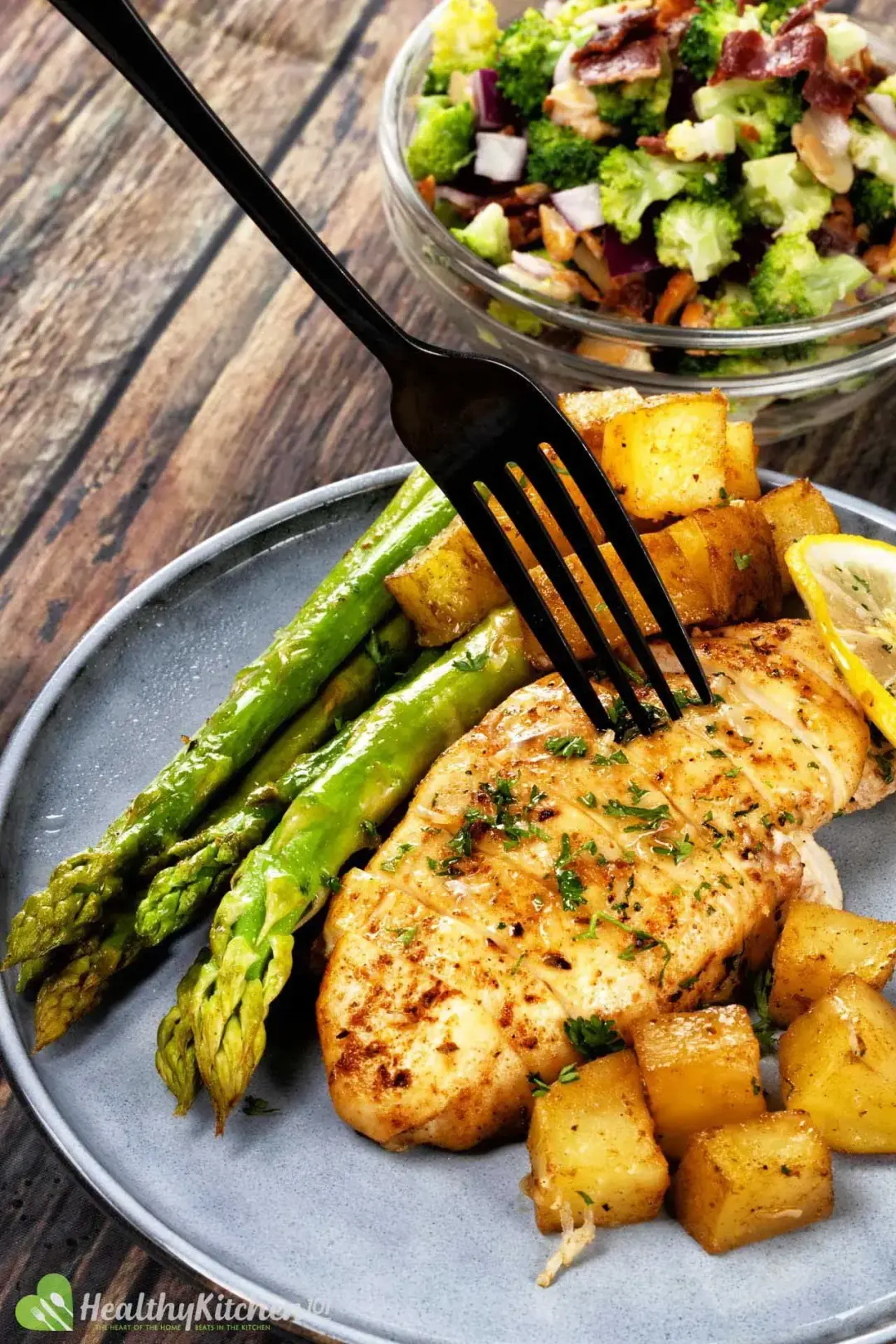 A black fork stuck into a piece of baked chicken breast surrounded by asparagus stalks and potato cubes