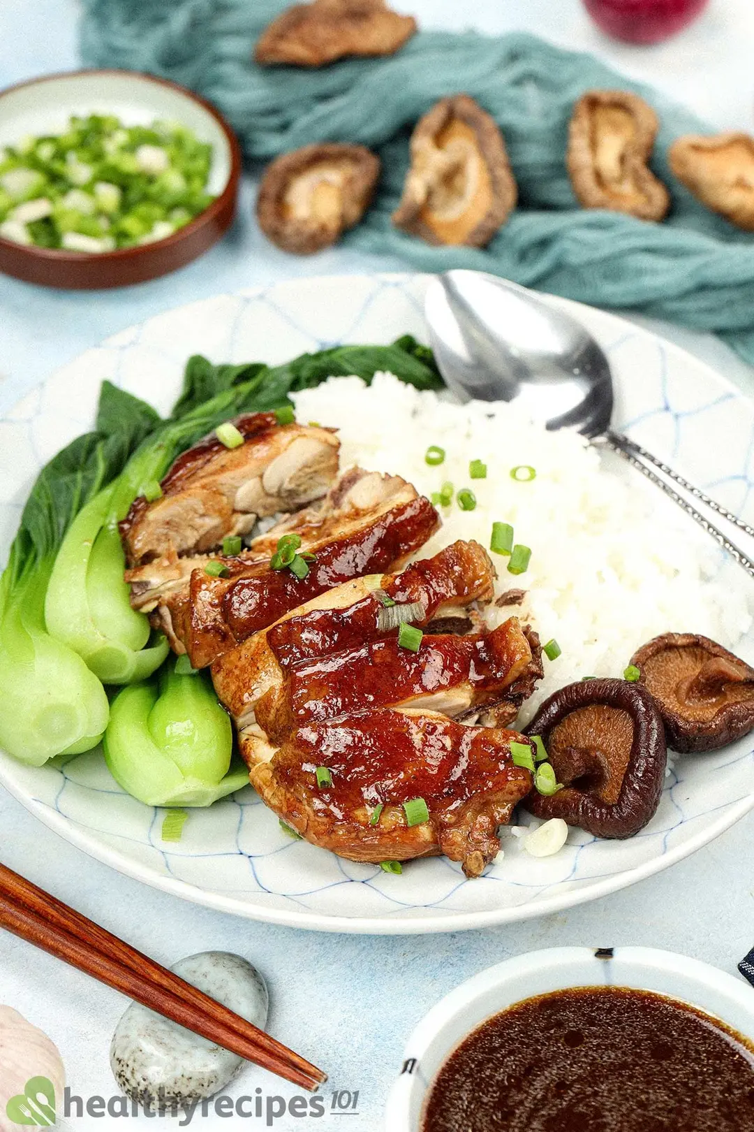 A plate of Asian food, containing rice, pieces of chicken, shiitake mushrooms, bok choy, chopped scallions, and a spoon