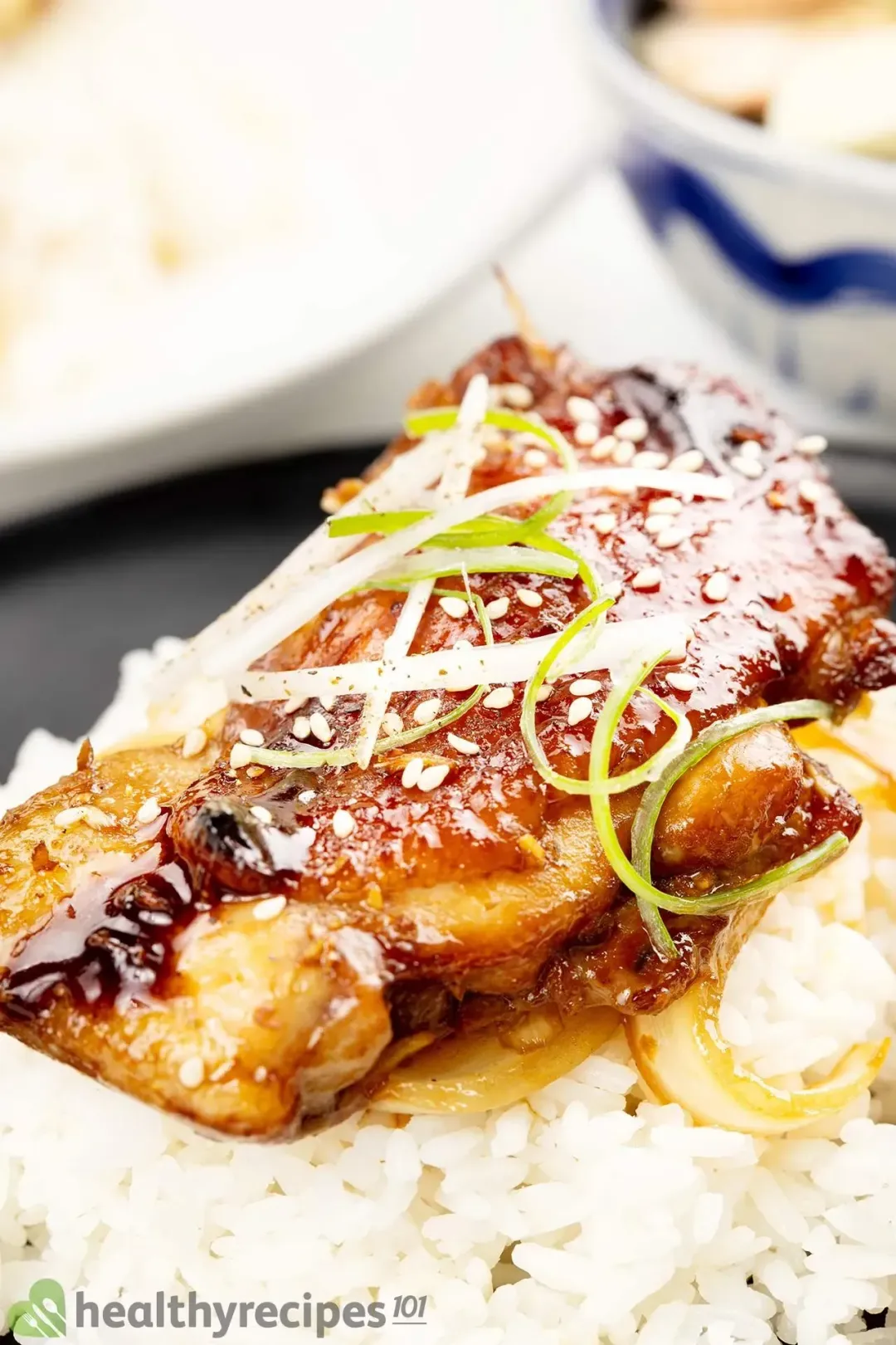 A close-up shot of teriyaki chicken and rice, with white sesame seeds, shaved green onion, and glossy sauced spooned on top
