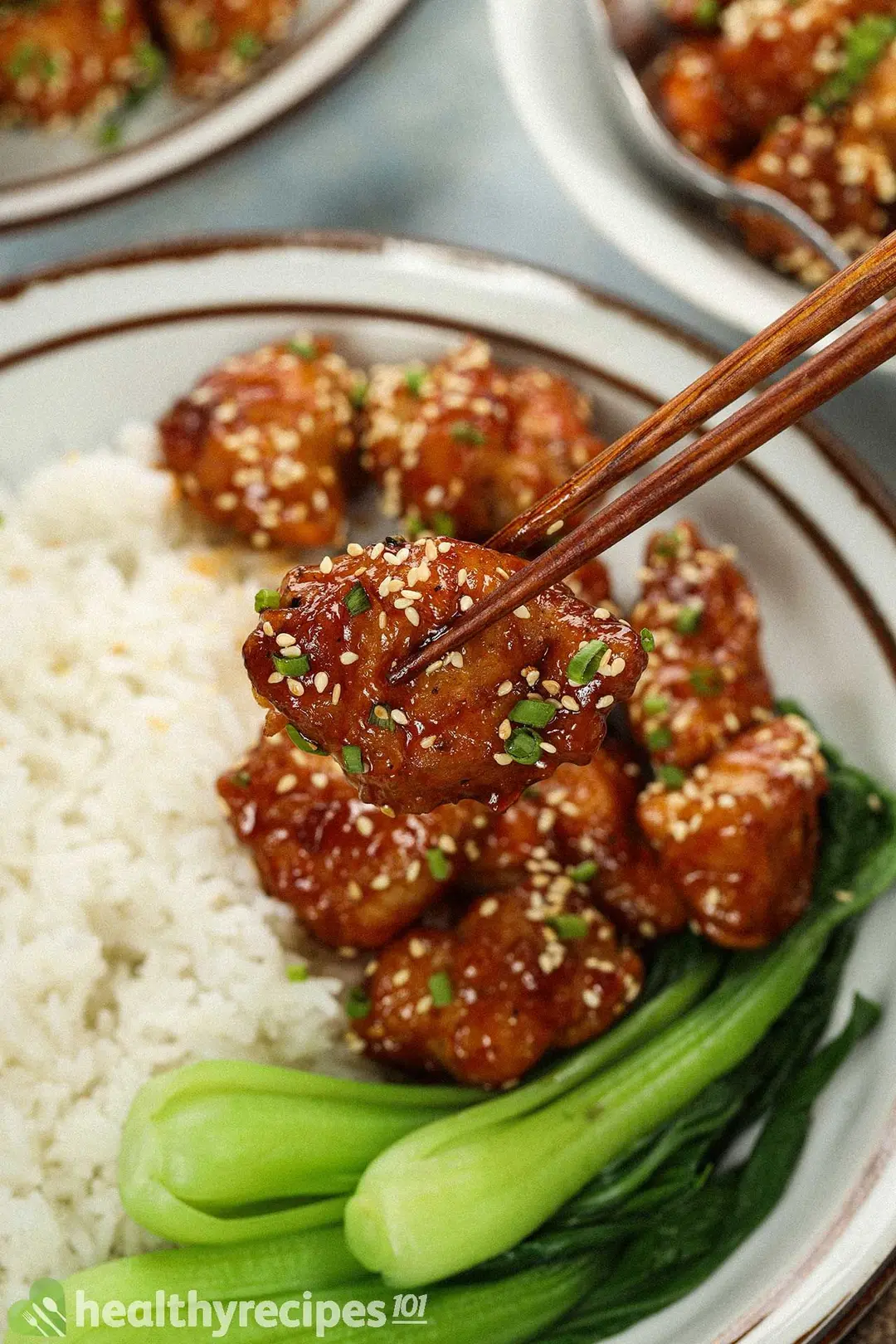 A pair of chopsticks holding up a piece of deep-fried chicken covered in honey sauce and white roasted sesame seeds