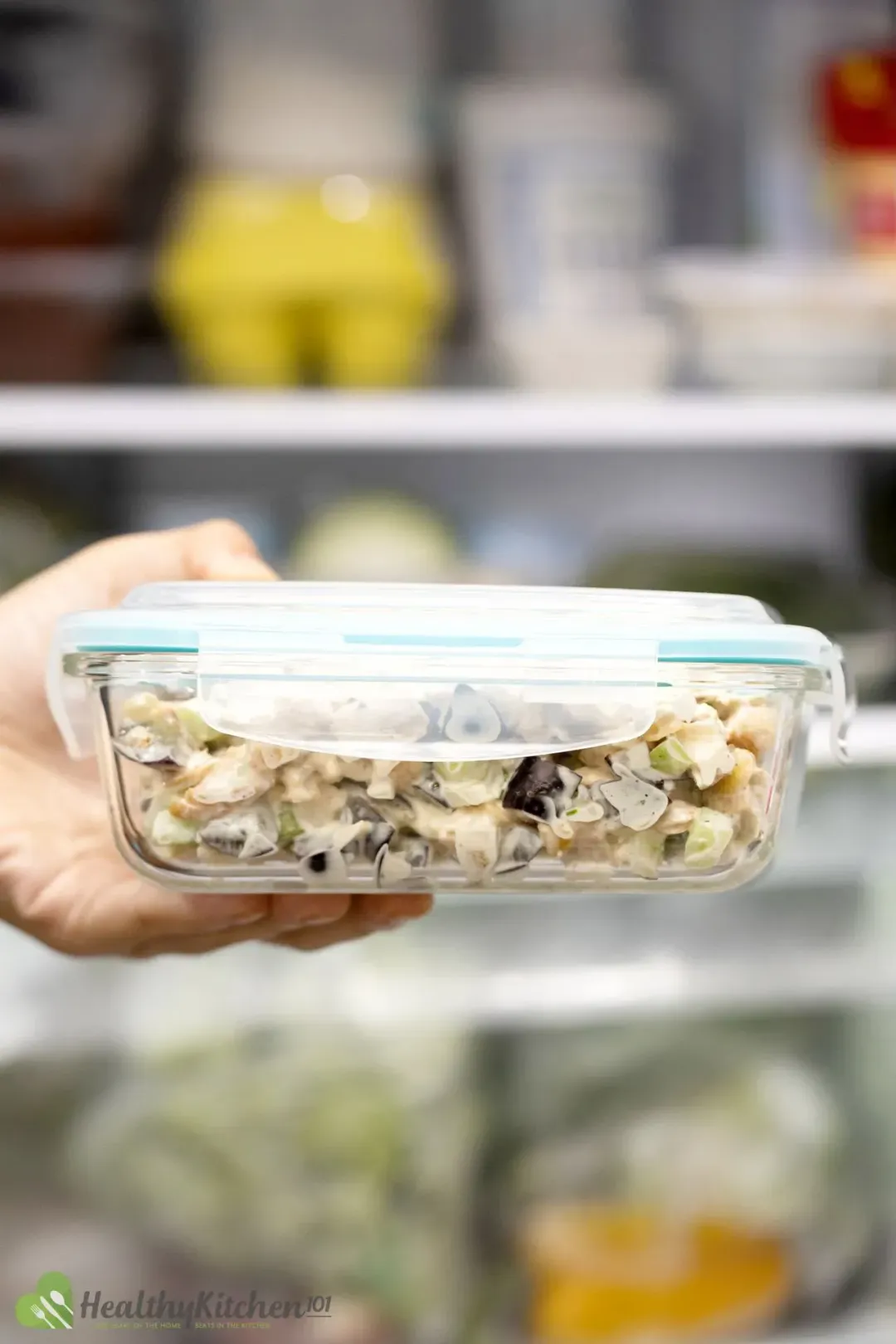 A hand holding a closed container of chicken and grape salad in front of an open fridge