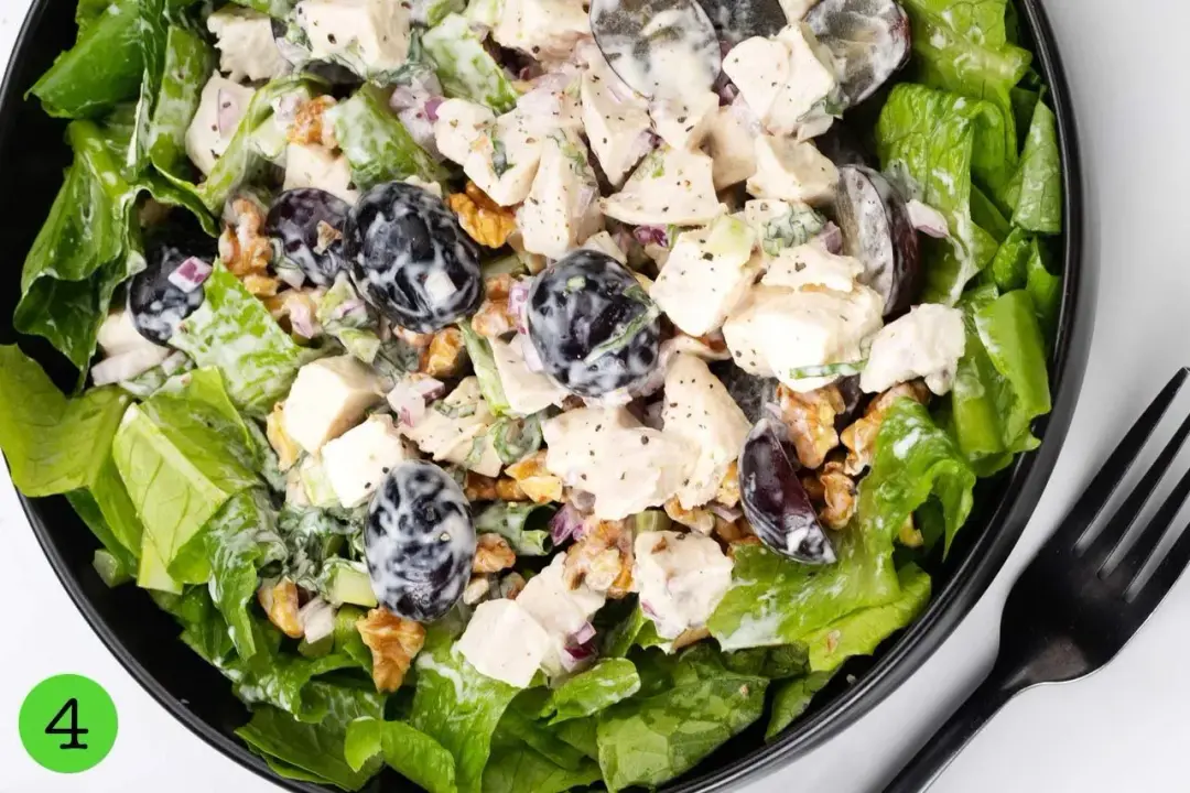 A black plate of waldorf salad with chopped lettuce, chicken breasts, walnuts, and dark grapes covered in mayonnaise dressing