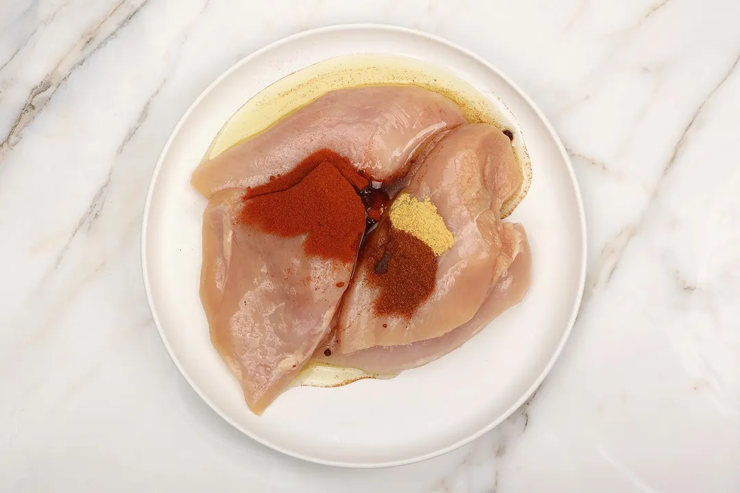 three chicken breasts with oil and seasoning on a white plate