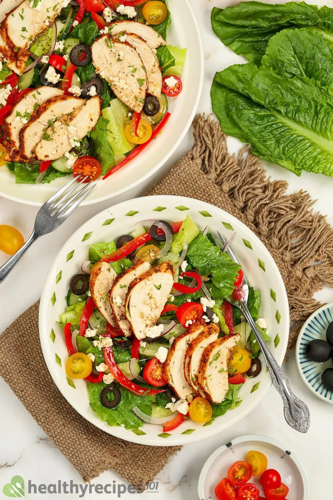 A high angle shot of two plates of greek chicken salad placed on a brown cloth and near cherry tomatoes, black olives, and a leafy green.