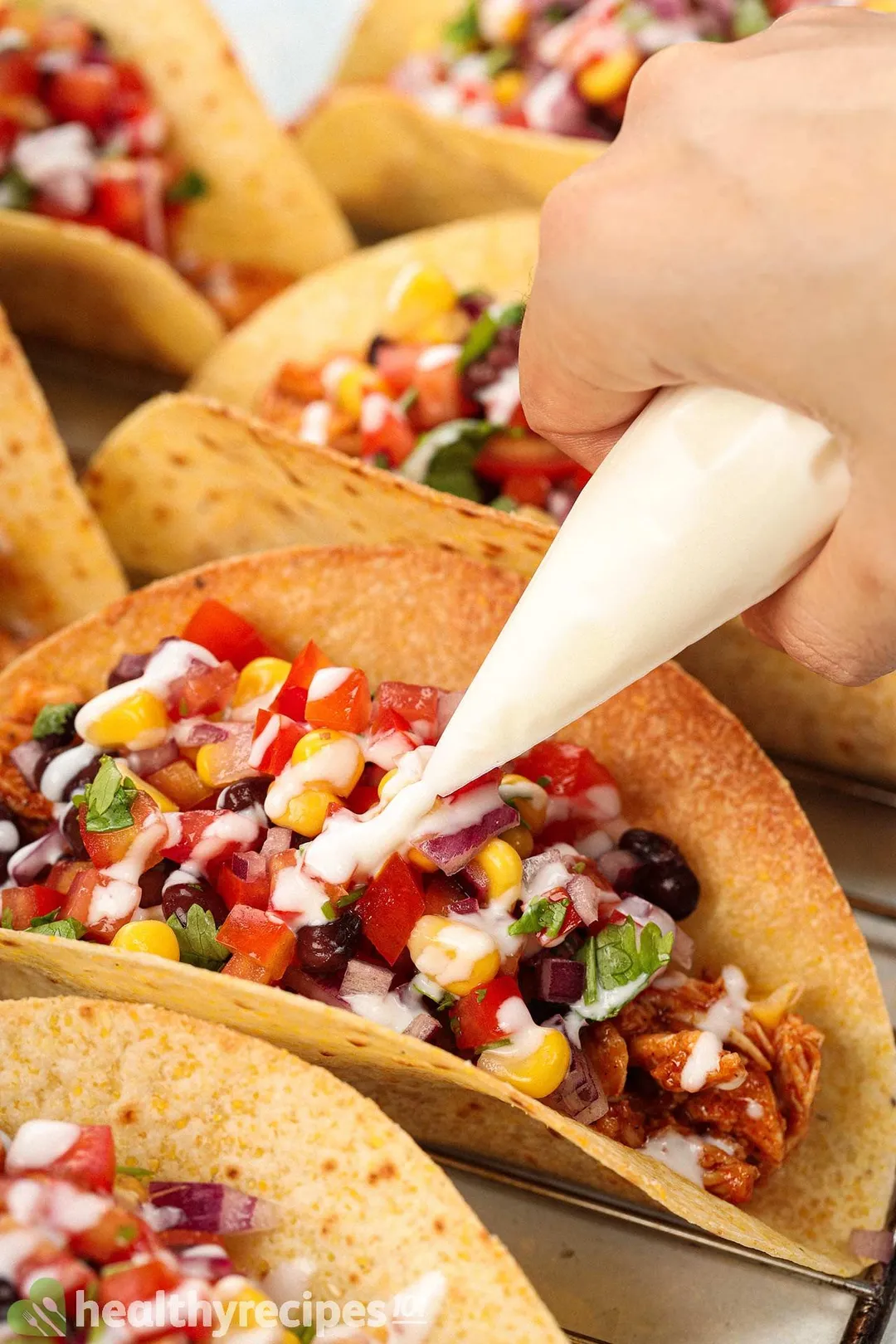 A hand drizzling yogurt with a piping bag onto BBQ chicken tacos