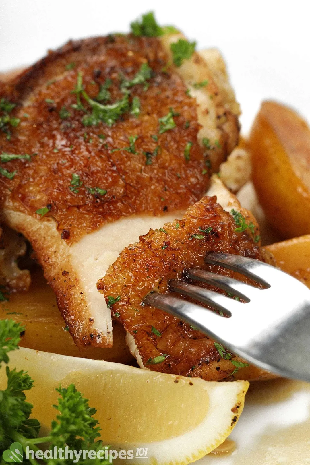 A close-up shot of a fork piercing into a piece of brown cooked chicken, laid near a lemon wedge and fresh parsley