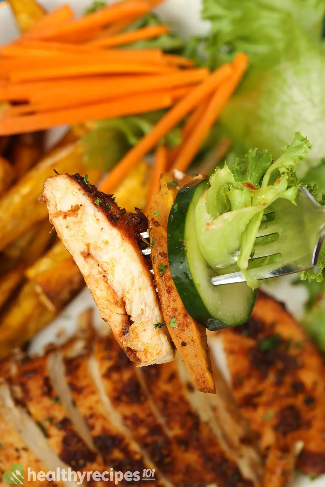 A fork sticking into a piece of chicken, a sweet potato fry, a cucumber slice, and a piece of lettuce