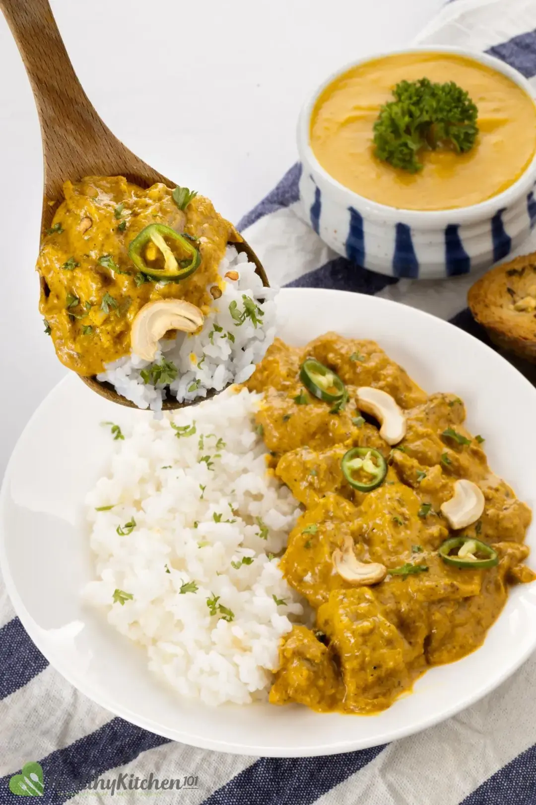 A plate of cooked rice and chicken korma, with a wooden spoon hovering over and a pumpkin soup in the background