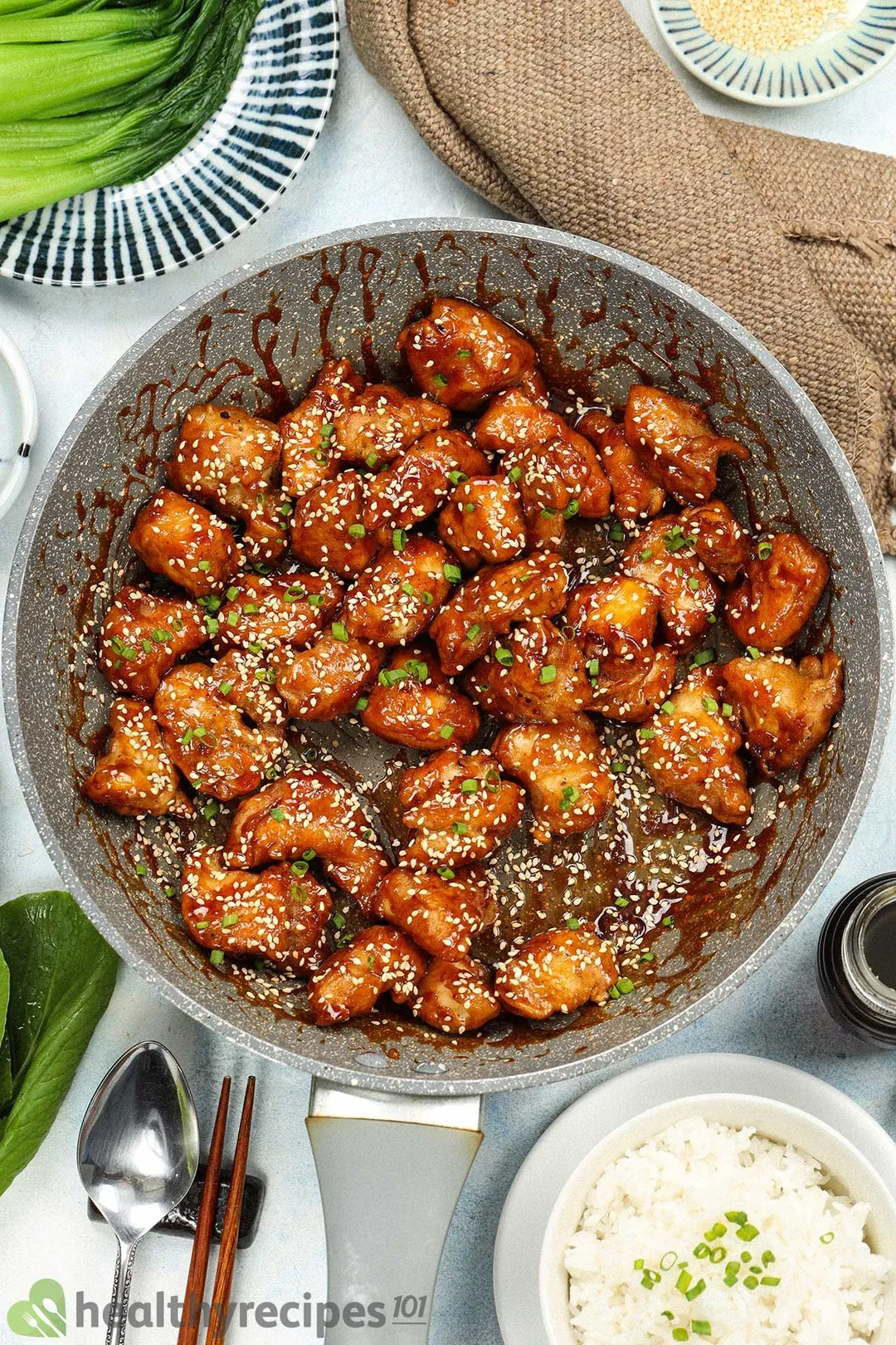 A large pan filled with pieces of honey chicken covered in roasted white sesame seeds and surrounded by a white rice bowl, a spoon, a pair of chopsticks, and a brown tablecloth