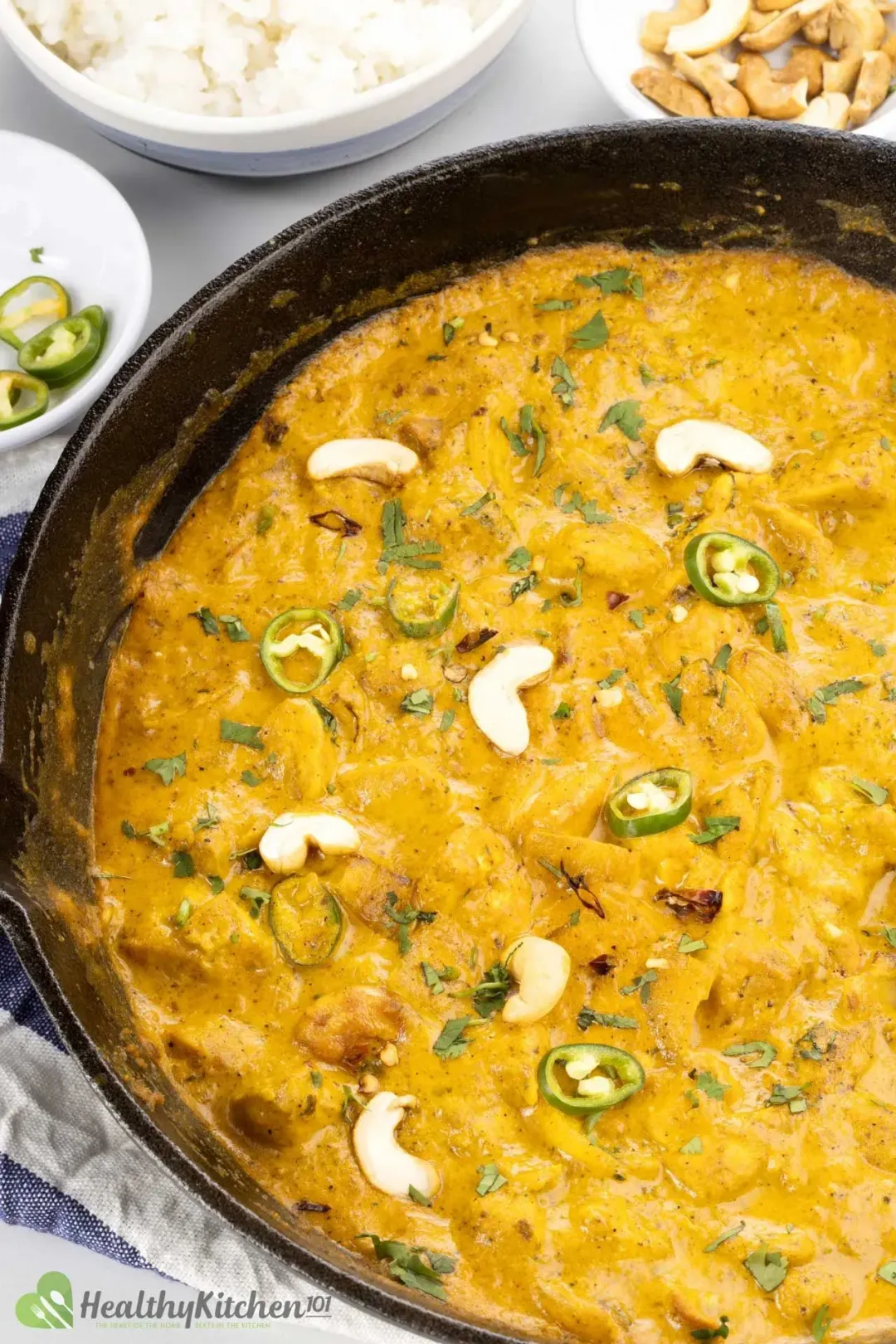 A black skillet of golden chicken korma topped with halved cashews and sliced jalapeños