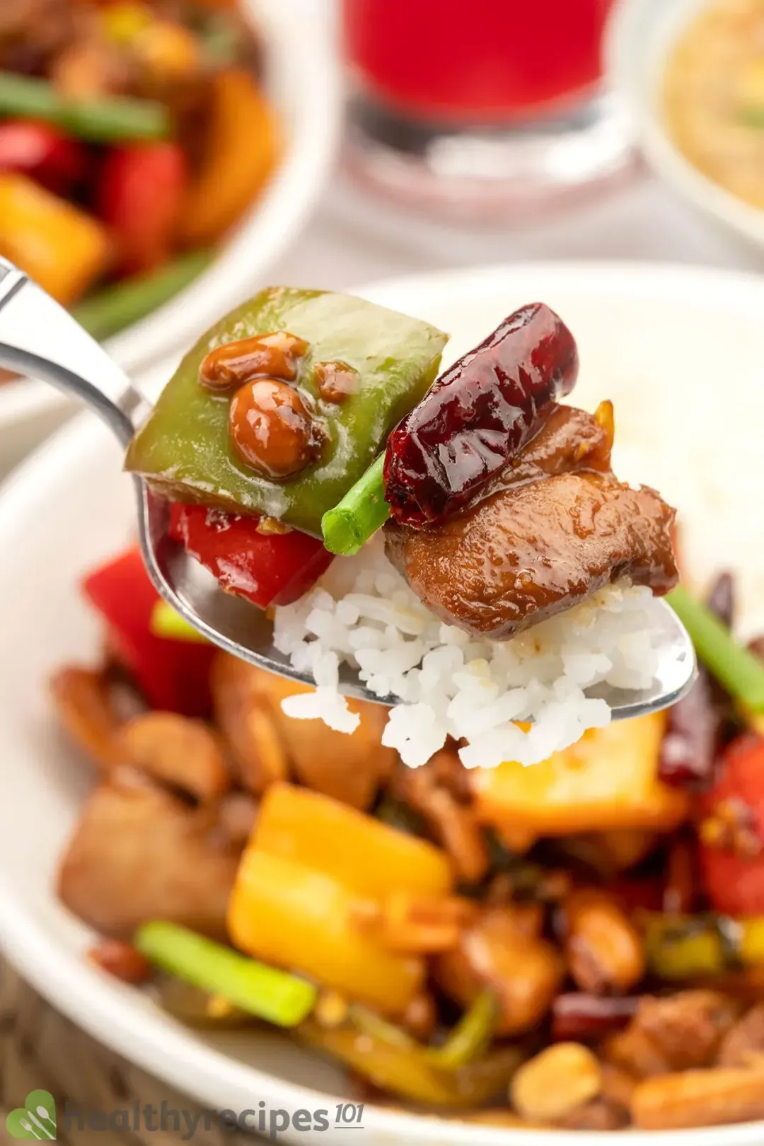 A spoon scooping up white rice, cicken cubes, dried chili, and diced bell pepper from a plate of kung pao chicken.