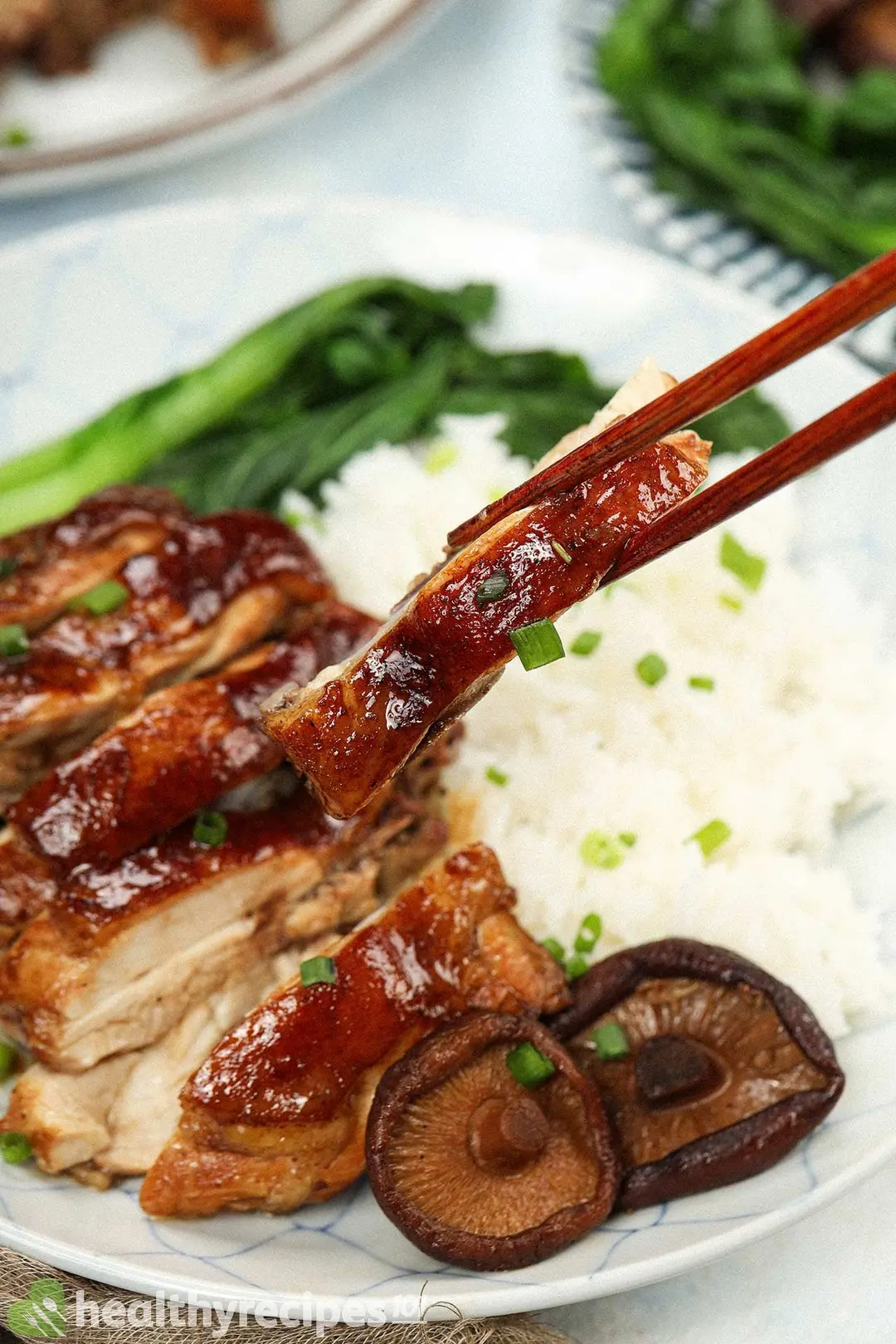 A pair of chopsticks picking up a piece of cooked chicken with a plate of rice, chicken, bok choy, and rice in the back