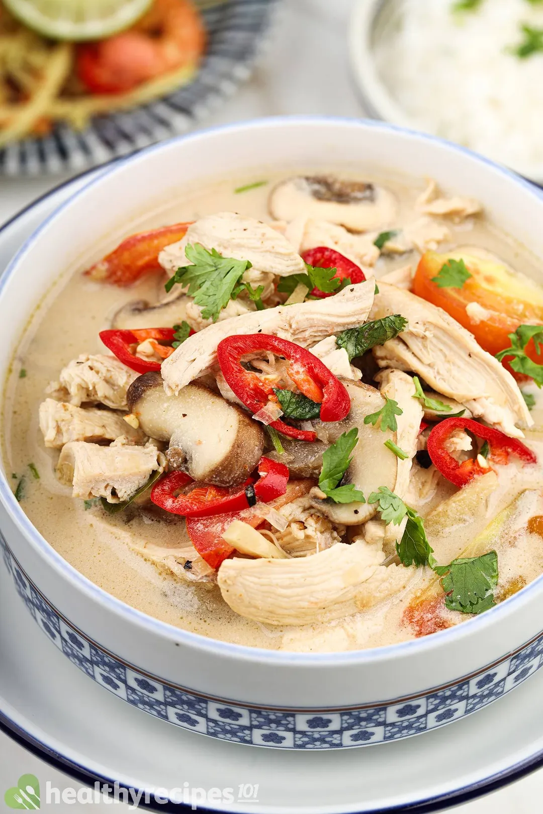 a bowl full of tom kha soup with shredded chicken, slices red jalapeno and mushroom on top