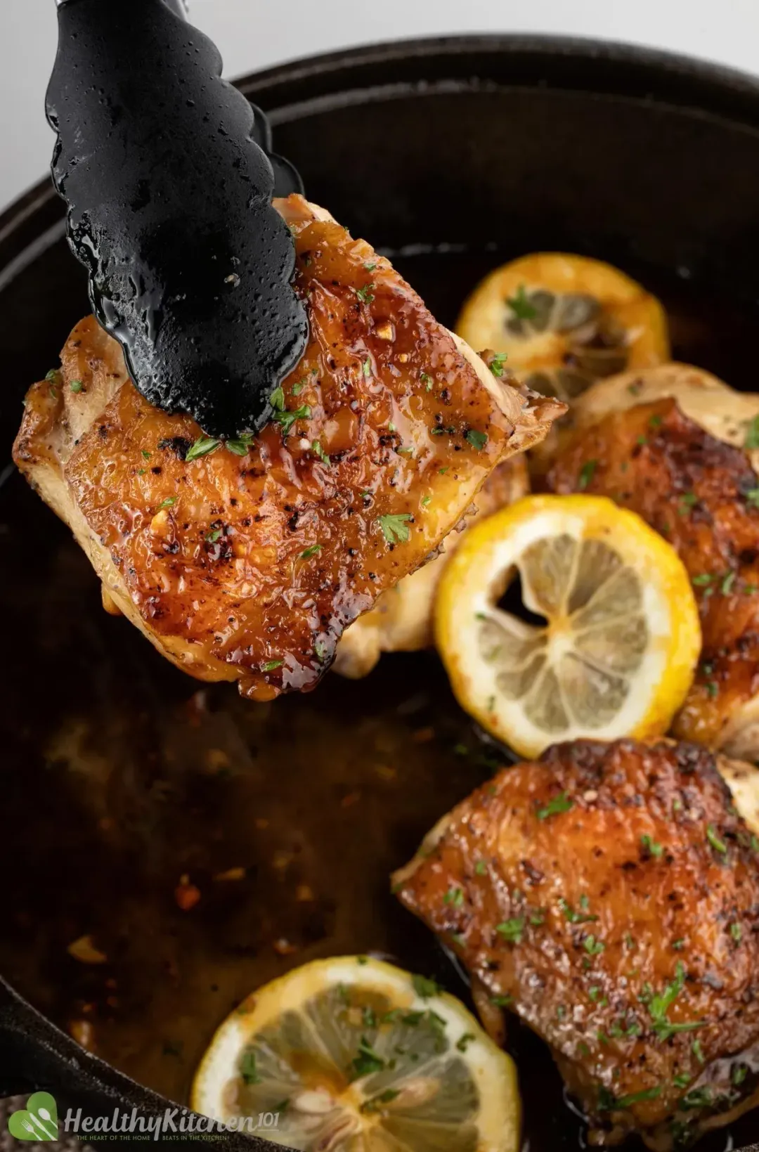 A pair of tongs holding a piece of golden chicken thigh over a skillet of golden chicken thighs and lemon wheels