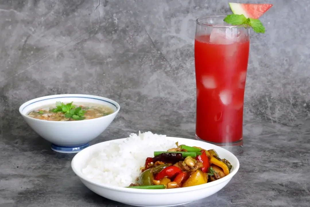 A plate of kung pao chicken and rice, a glass of watermelon juice, and a bowl of egg drop soup.