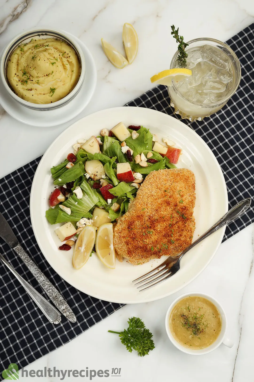 a plate of cooked chicken with salad next to a glass of lemon juice, a bowl of mashed potato and a cup of sauce