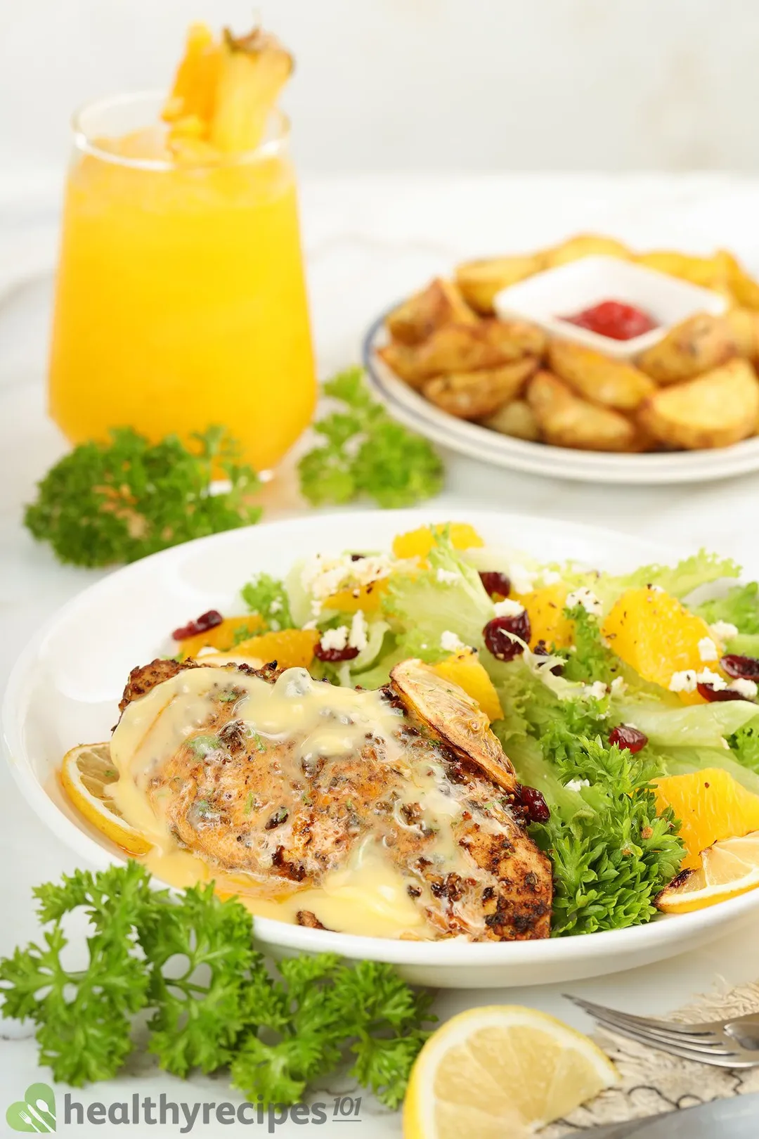 a plate of cooked chicken breast and salad decorated with a glass of orange juice and a plate of potato wedge