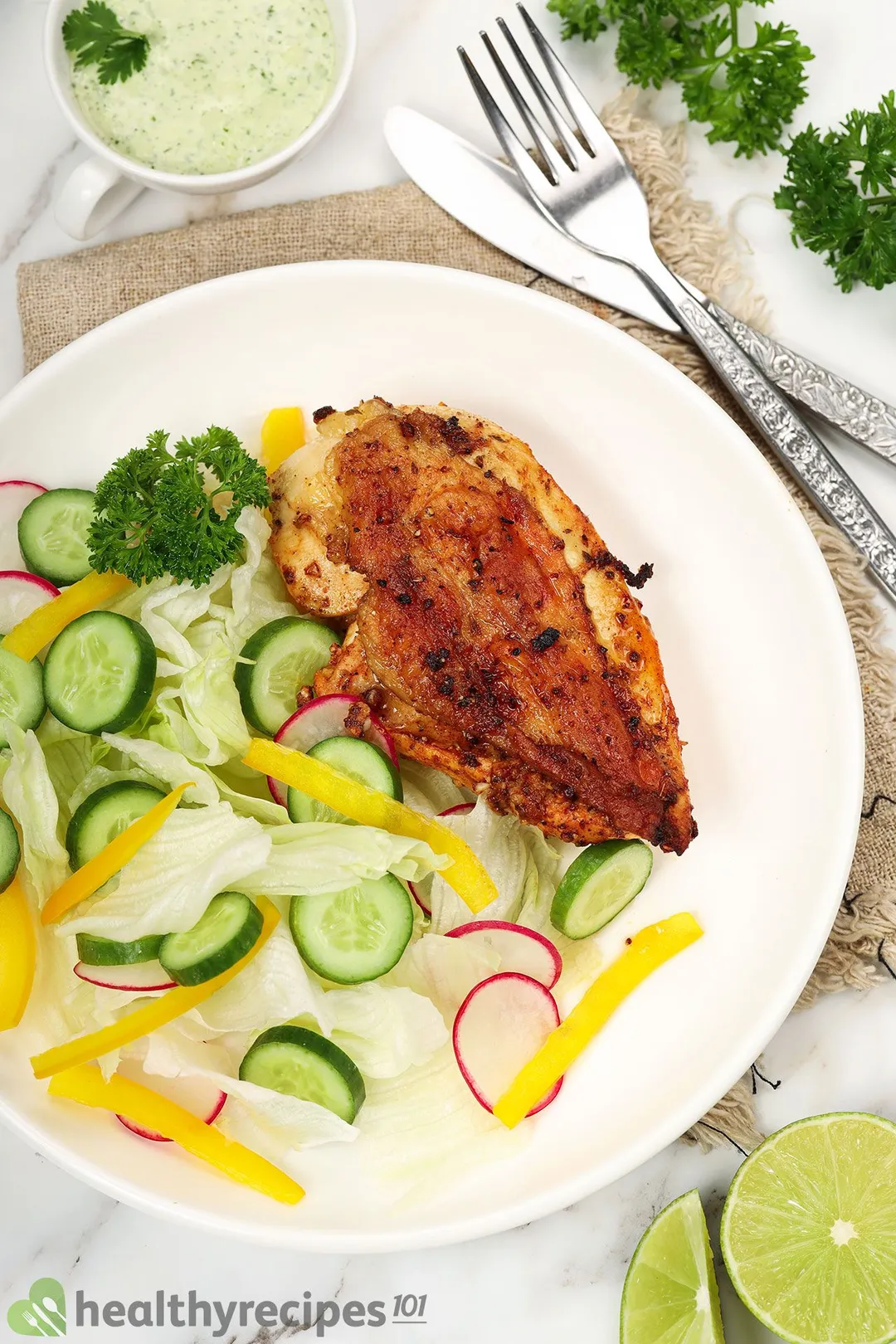 a plate of cooked chicken breast with sliced cucumber and chopped lettuce decorated with half lime, spoon and fork and a small bow of sauce