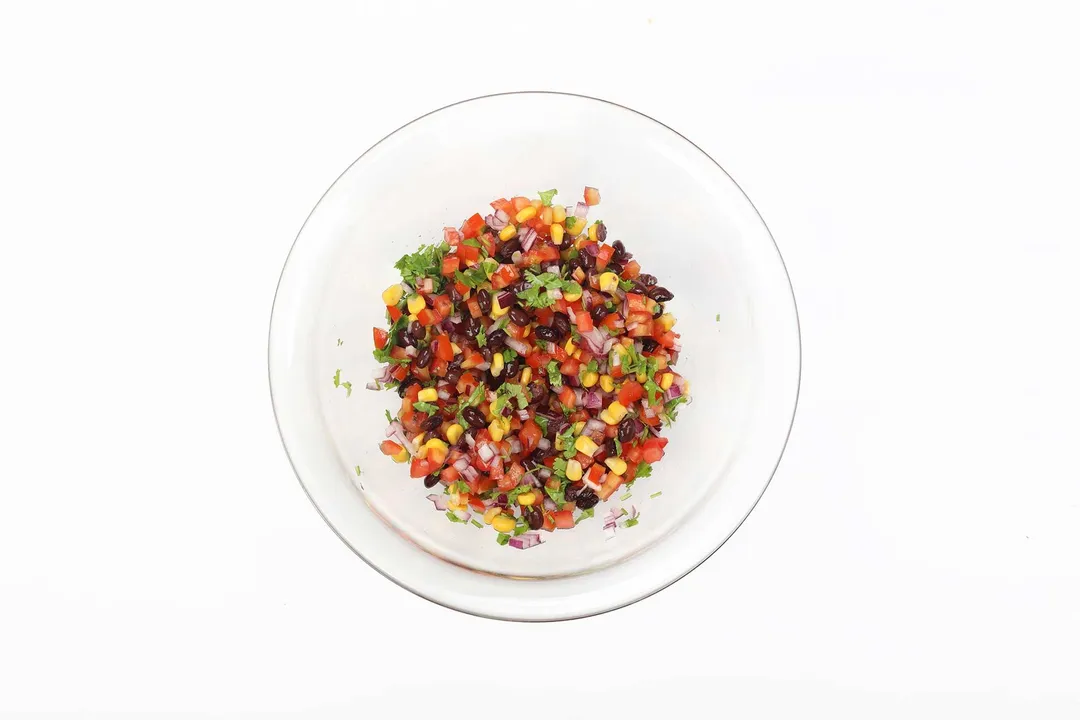 A large glass bowl containing a colorful mixture of diced tomatoes, red beans, corn, and chopped parsley