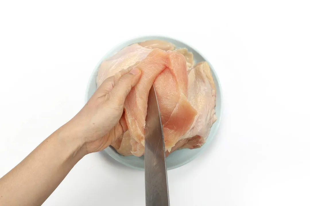 A hand holding a raw chicken thigh while a knife is making a slit into it