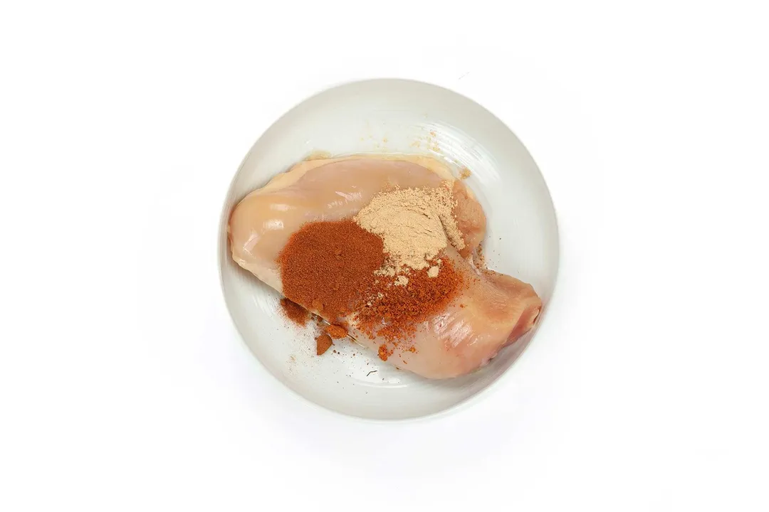 Two pieces of raw chicken breasts laid on a white plate with a large amount of paprika and garlic powder laid over top