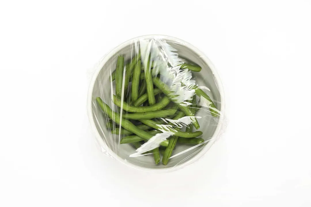 A bowl containing green beans covered in cling wrap