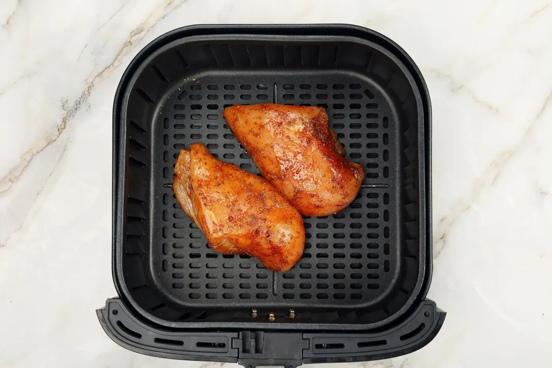 Two uncooked chicken breasts covered in spices placed inside an air fryer basket