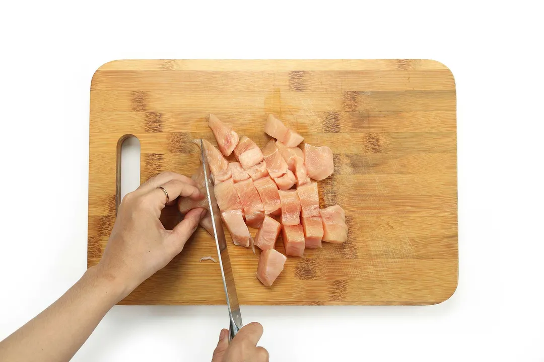 A hand holding a knife and slicing raw chicken into small cubes on a rectangle wooden chopping board