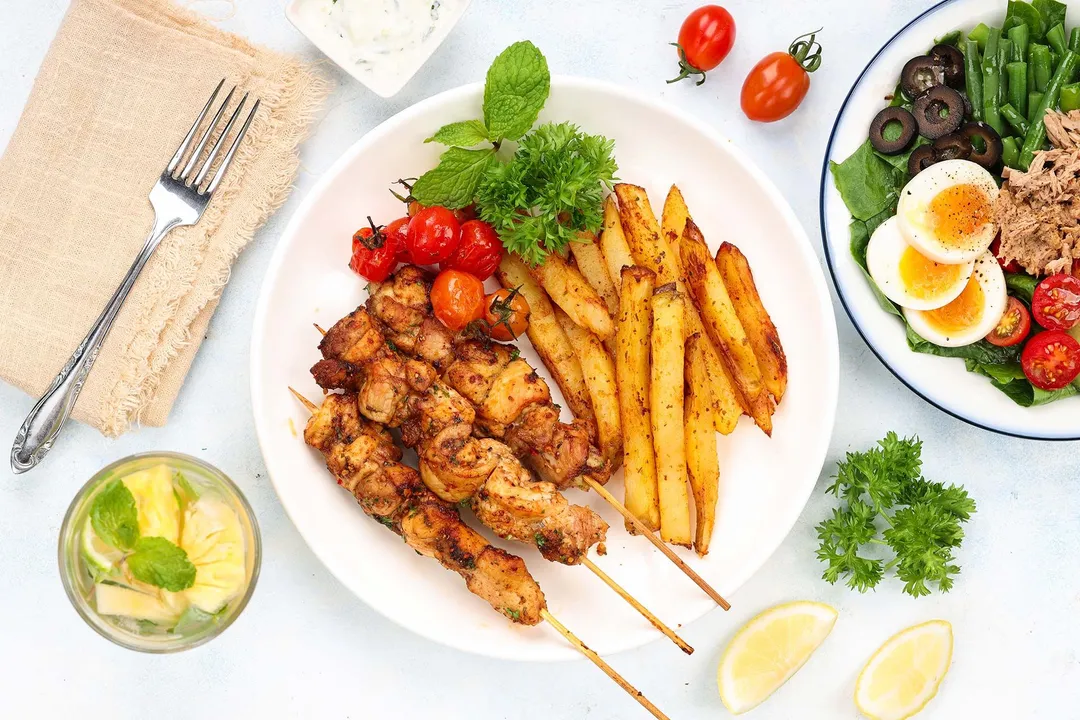 a plate of cooked chicken, potato and tomato decorated with a plate of salad and a glass of water