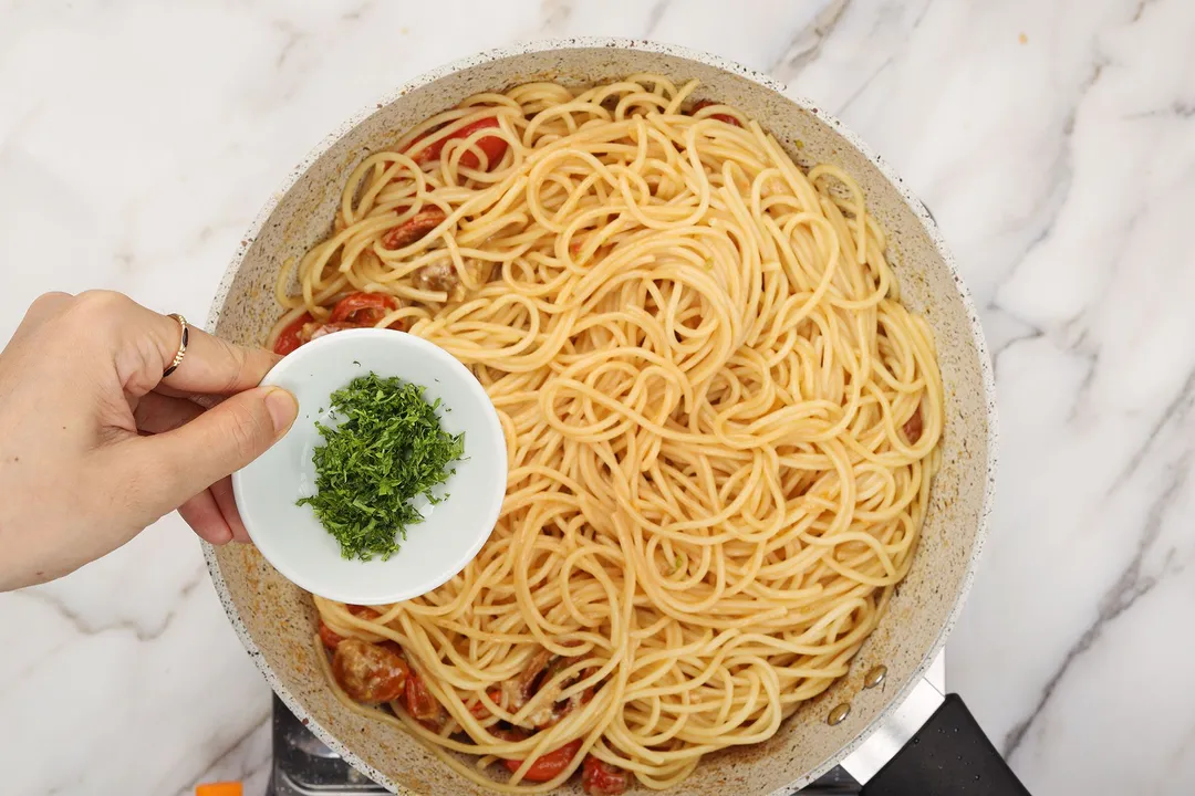 hand holding a small bowl of chopped parsley on top of a skillet of pasta