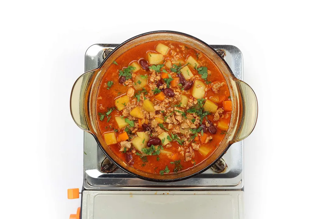 A glass saucepan cooking ground chicken soup filled with cubed potatoes, ground chicken, coarsely diced carrots, and chopped herbs