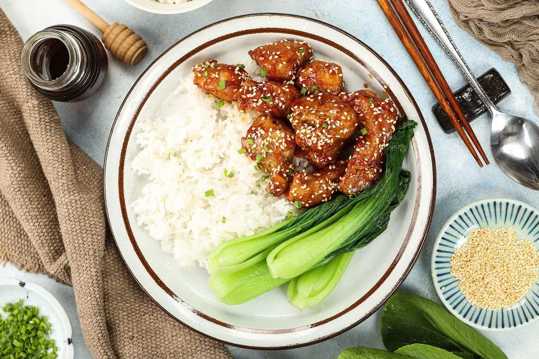 A high-angle shot of a honey chicken plate featuring pieces of honey chicken, white bowl, and bok choy; a brown tablecloth, a small plate of white sesame, a spoon, chopsticks, and a honey pot