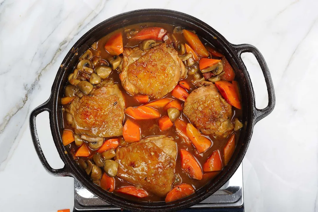 a cast iron skillet of cooked chicken thighs, chopped carrot, mushroom and sauce