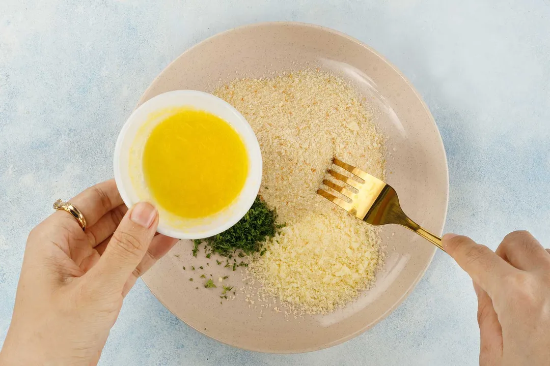 a hand holding a small bowl of yellow sauce and another hand holding a fork on a plate of plain panko breadcrumbs and chopped parsley