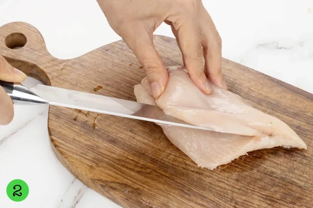 A hand using a knife to butterfly cut a raw chicken breast placed on a wooden chopping board.