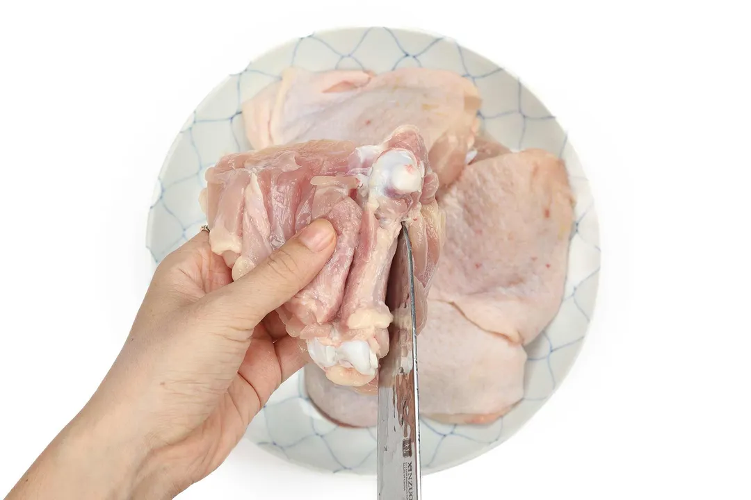 A hand holding a raw chicken thigh with a knife making a small slit into it