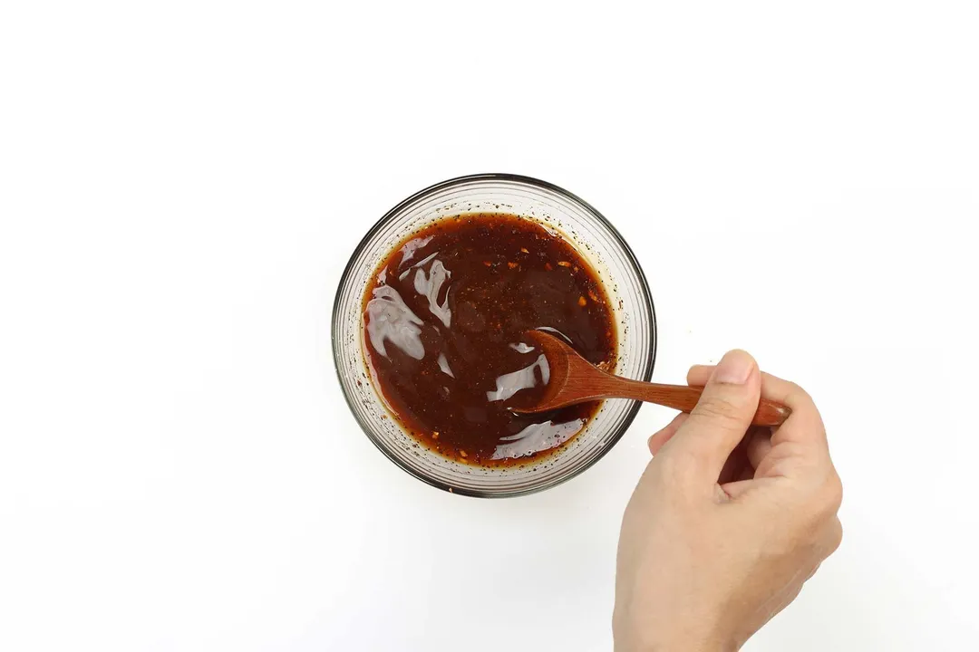 A hand stirring a dark red and glossy sauce in a sauce bowl with a wooden spoon
