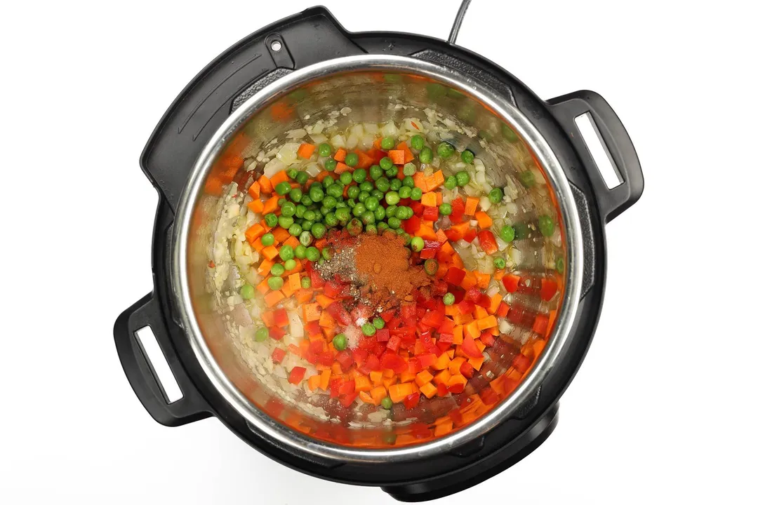 An instant pot's inner pot containing diced bell peppers, diced carrots, green peas, and spices