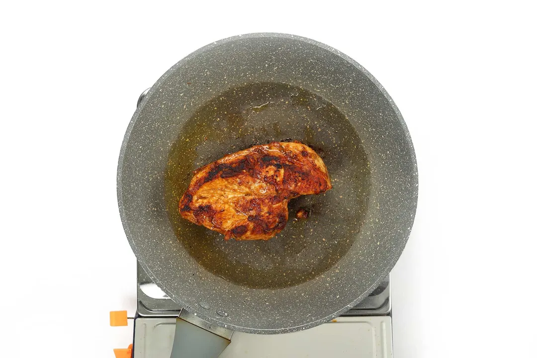 A large pan cooking a piece of chicken breast whose exterior has turned golden with black charred marks
