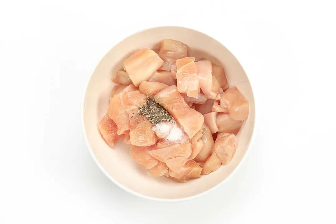 A white bowl containing raw chicken cubes with a small amount of salt and black pepper in the center
