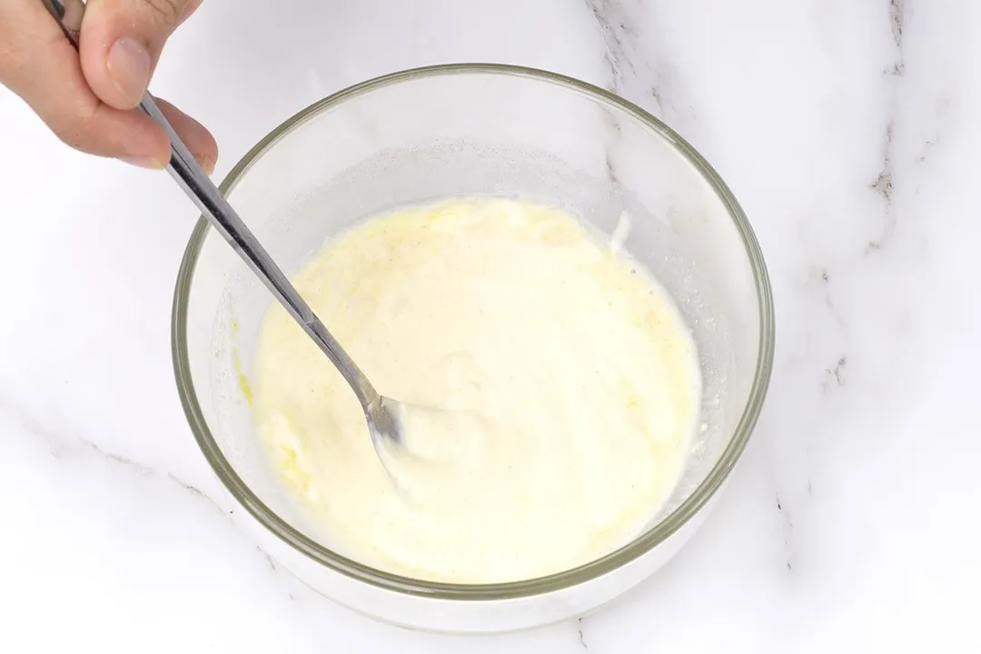 a hand holding a spoon to whisk white sauce in a bowl