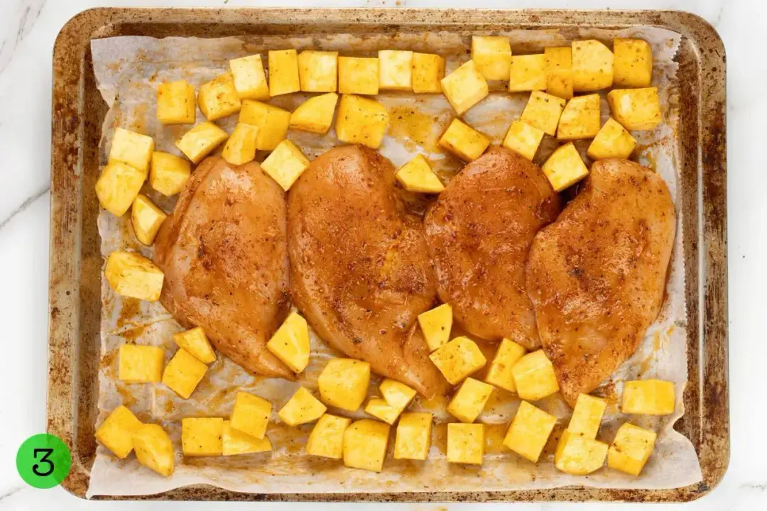 Four pieces of seasoned chicken breasts and numerous potato cubes laid on a baking pan lined with parchment sheet