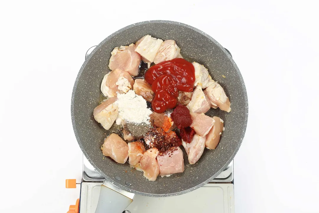 A large pan cooking large raw chicken cubes with dollops of ketchup and several spices laid in the center