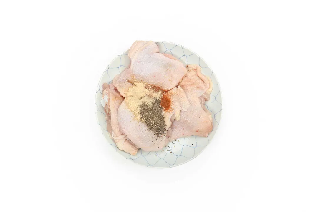A round plate containing four raw chicken thighs with black pepper, garlic powder, and paprika in the center