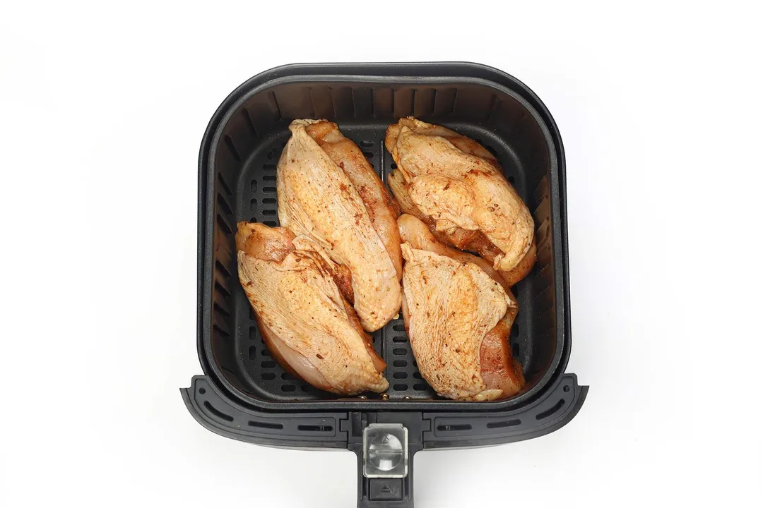 An air fryer basket containing four raw chicken thighs covered in paprika