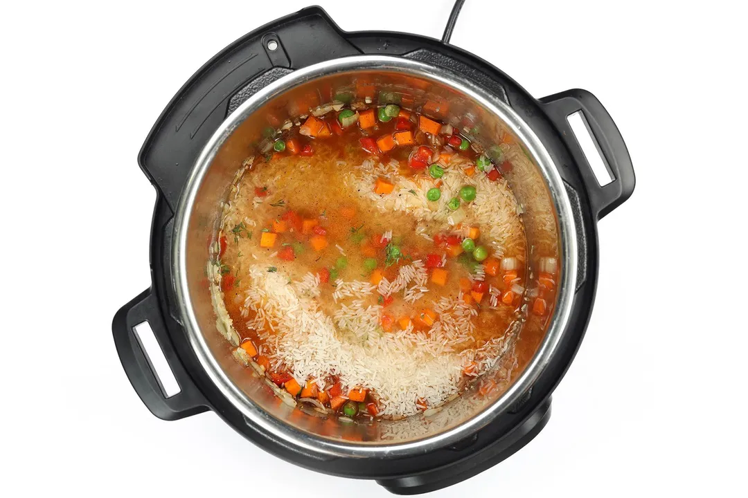 An instant pot's inner pot containing diced carrots, diced bell peppers, green peas, rice, and chicken broth
