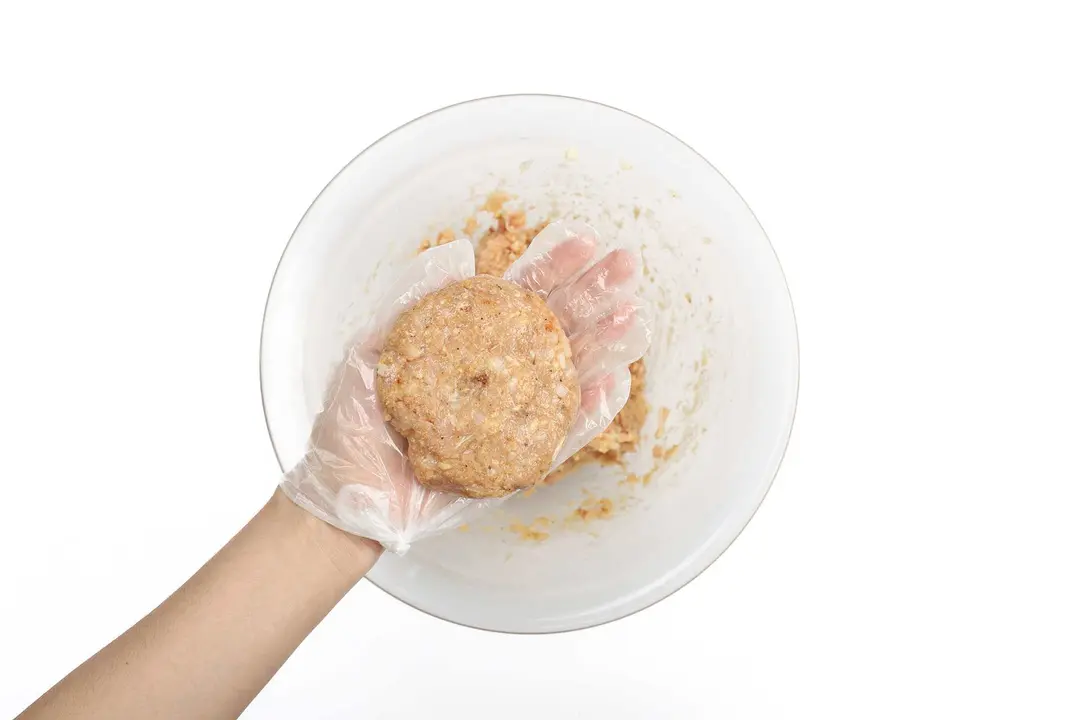 A gloved hand holding a meat pattie over a large glass bowl