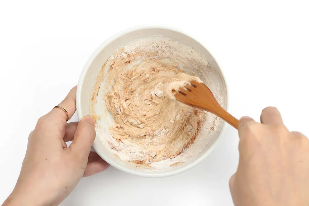 A hand mixing a thick and beige batter in a white bowl with a wooden fork